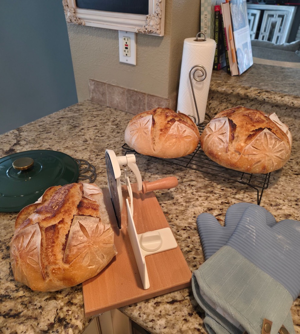 Scored sourdough loaves next to a bread slicer and oven mitts